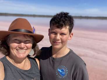 A woman in a brown hat and a boy smile together, standing close, with a pink-hued lake and clear blue sky in the scenic background.