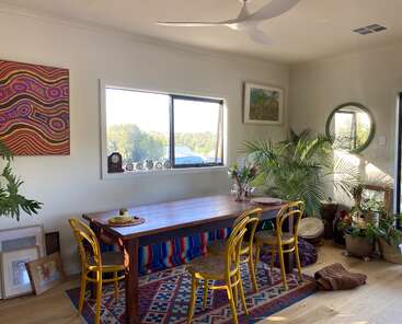 A bright dining room features a wooden table with yellow chairs, vibrant rugs, green houseplants, colorful wall art, a window view, and decorative ceramics. Relaxed, inviting atmosphere.