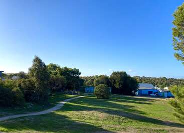 A sunny, clear day with vibrant blue sky, green grass, and scattered trees. A winding path leads to small blue buildings nestled among the foliage.