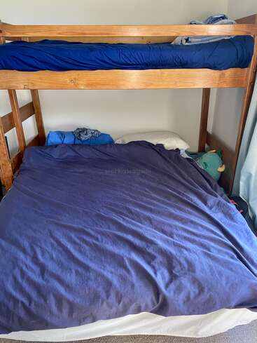 This image shows a wooden bunk bed with blue bedding. The top bunk is neatly made, while the bottom bunk appears larger and slightly messy with pillows.
