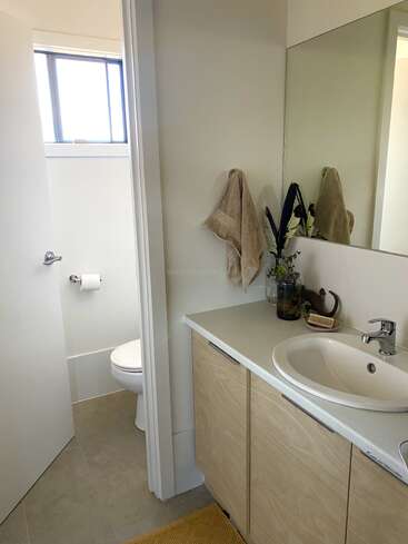 This image shows a modern bathroom with a white sink, light wooden cabinets, a mirror, beige towels, a plant, and a separate toilet area with a window.