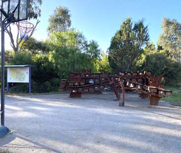 The image shows a park area with unique, rust-colored metal benches shaped like branches, surrounded by trees. A basketball hoop and information board are visible.