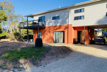 A modern two-story house with orange lower walls, white upper siding, a balcony with glass railing, large windows, sliding doors, and a gravel driveway.