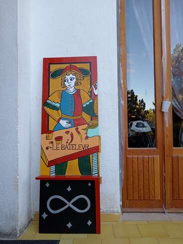 Dieses Bild zeigt ein farbenfrohes Schild mit der Tarotkarte "Le Bateleur", das an einer weißen Wand in der Nähe einer Holztür steht, mit einem Unendlichkeitssymbol darunter.