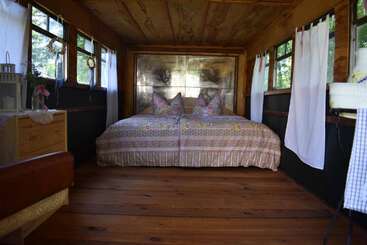 This cozy room features a large bed with patterned bedding, wooden floors, windows with white curtains, rustic decor, and natural light creating a warm, inviting atmosphere.