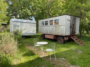 Two old trailers sit in a green, sunny yard surrounded by trees. In front, a small white table and two chairs create a cozy outdoor setting.