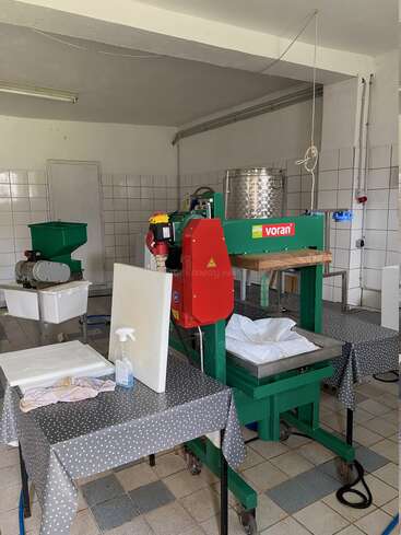 The image shows a small, tiled processing room with a green and red Voran fruit press, stainless steel tank, tables with cloths, and cleaning supplies. Industrial, clean environment.