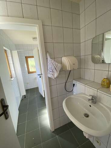 The image shows a tiled bathroom with a white sink, mirror, soap dispenser, towel, and a partially open door leading to another bright, tiled room.