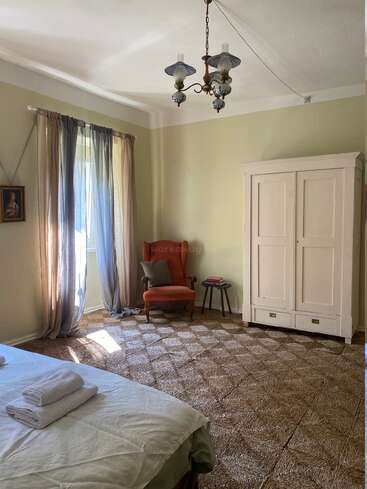 This cozy bedroom features a vintage chandelier, large wardrobe, red armchair, small side table with books, textured carpet, bed with towels, and sheer curtains.
