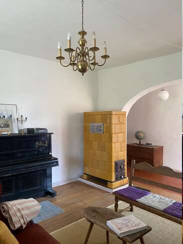 A cozy vintage living room with a brass chandelier, black piano, yellow tiled stove, wooden furniture, globe, book, and rustic decor creating a warm atmosphere.