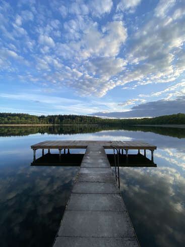 A tranquil lake reflects the dramatic cloudy blue sky. A wooden pier stretches forward, surrounded by calm water and lush green forest on the distant shore.