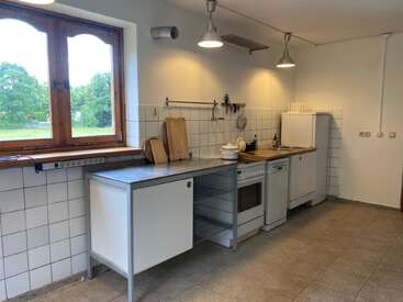 A minimalist kitchen with white tiled walls, stainless steel counters, wooden cutting boards, white appliances, and large window overlooking a green landscape, brightly lit.
