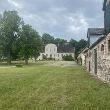 A large, old building stands in the background surrounded by trees. Stone outbuildings line the right side, with a grassy courtyard under a cloudy sky.