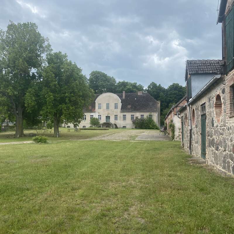 A large, old building stands in the background surrounded by trees. Stone outbuildings line the right side, with a grassy courtyard under a cloudy sky.