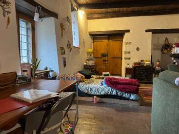 A cozy, rustic living space with wooden ceiling beams, a low bed, colorful bedding, books, a wooden table, houseplants, and eclectic decorations scattered throughout the room.