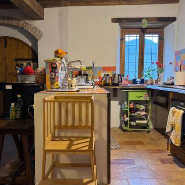 This cozy kitchen features rustic wooden beams, a wooden high chair, colorful tiles, a sunny window, various appliances, plants, and shelves full of household items.