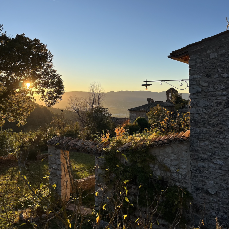 Golden sunlight bathes a rustic stone house and lush garden, casting long shadows. Mountains and a clear sky create a tranquil, picturesque countryside sunset scene.