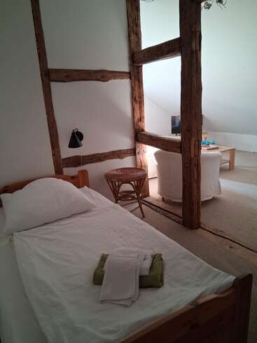 Una acogedora habitación en el ático cuenta con una cama individual con sábanas blancas, vigas de madera rústicas, una mesa de mimbre, dos sillones y un ambiente luminoso y relajado.