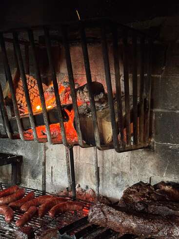 Una parrilla rústica con brasas encendidas y leños ardiendo arriba, salchichas y filetes chisporroteando en la rejilla metálica abajo, creando una escena acogedora y humeante de barbacoa.