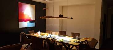 A modern dining room with a long wooden table, brown chairs, abstract wall art, and a linear pendant light. Some items and jackets are on the chairs.