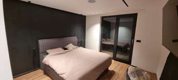 This modern bedroom features a large bed with light bedding, black accent wall, wooden floor, glass doors, minimal decor, and soft lighting, creating a cozy atmosphere.