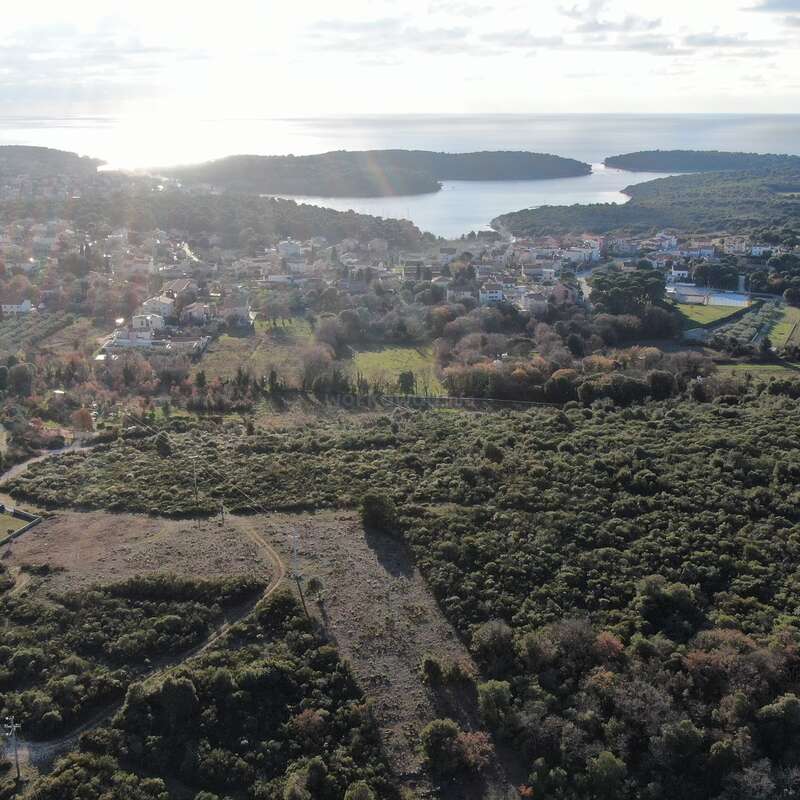 This aerial image shows a lush, green landscape with scattered houses, winding roads, dense forest, and a coastal town beside calm blue water under a bright sky.