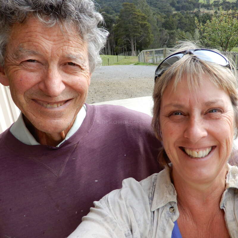 A imagem mostra uma selfie de um homem e uma mulher mais velhos em pé do lado de fora, com uma estrada de cascalho, árvores e uma cerca ao fundo.