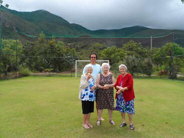Vier ältere Frauen und eine jüngere Person stehen auf einem grasbewachsenen Feld mit einem Fußballtor, umgeben von Netzen und Bergen unter einem bewölkten Himmel.