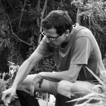 Un homme portant des lunettes et un t-shirt est assis à l'extérieur, penché en avant, peut-être en train de travailler dans un jardin. La photo en noir et blanc montre des plantes et des arbres autour de lui.