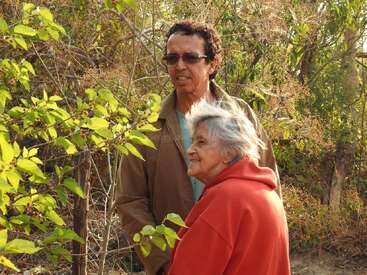 Eine ältere Frau in einer roten Jacke steht lächelnd neben einem Mann mit Brille und einem braunen Mantel, umgeben von grünem Laub in einer natürlichen Umgebung im Freien.
