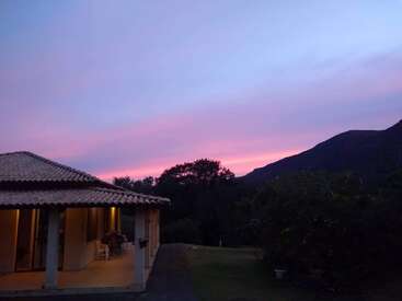 Ein friedliches Haus mit warmen Lichtern leuchtet in der Abenddämmerung. Der Himmel ist wunderschön in Violett- und Rosatönen gemalt, mit Hügeln und Bäumen, die sich als Silhouetten abzeichnen.