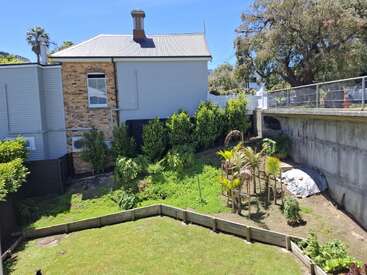Une arrière-cour ensoleillée comprend une petite pelouse bordée d'une clôture en bois, de jeunes arbres, des buissons luxuriants, une vieille maison en briques et un mur de soutènement en béton.