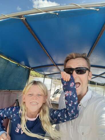 Ein Mann und ein junges Mädchen lächeln und posieren spielerisch für ein Selfie auf einem Boot mit einem blauen Vordach und genießen ein sonniges Abenteuer im Freien.