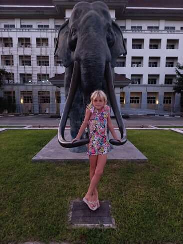 Ein junges Mädchen in einem bunten Kleid steht zwischen den Stoßzähnen einer großen Elefantenstatue und posiert spielerisch vor einem modernen Gebäude. Abendlicht.