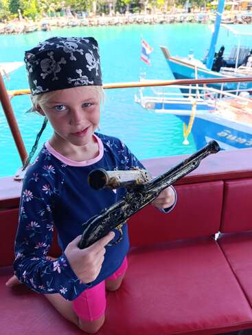 Ein junges Mädchen, das als Pirat gekleidet ist und ein Totenkopftuch trägt, hält eine Spielzeugpistole auf einem roten Sitz. Dahinter sind blaues Wasser und Boote zu sehen.