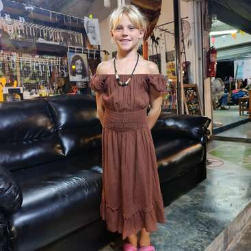 Ein lächelndes Kind in einem braunen schulterfreien Kleid und rosa Hausschuhen steht vor einem schwarzen Sofa in einem gemütlichen Künstleratelier oder einer Galerie.