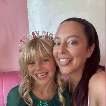 A smiling woman and young girl take a playful selfie together. The girl wears a "Birthday Girl" headband and green dress, celebrating a special birthday occasion.