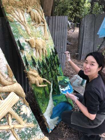 Une femme peint un grand panneau d'art extérieur texturé représentant des animaux, des arbres et une rivière. Elle sourit à l'appareil photo, tenant une palette de peinture.