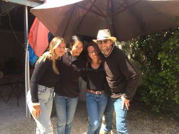 Cuatro personas permanecen juntas, sonriendo al aire libre bajo una gran sombrilla. Están relajados, llevan vaqueros informales y camisetas oscuras, y disfrutan de un día soleado con vegetación detrás.