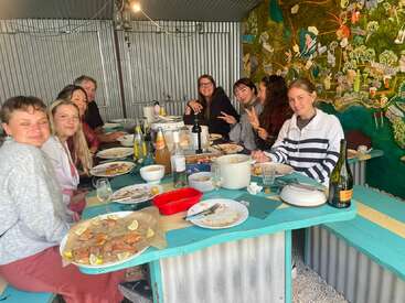 A group of friends enjoy a casual meal together at a vibrant turquoise table, surrounded by colorful wall art, smiling and sharing food in a cozy setting.