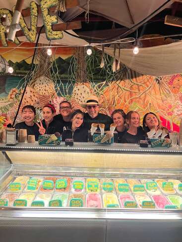 A cheerful group of people stand behind an ice cream counter, displaying a variety of colorful flavors. Festive decorations and a vibrant mural create a lively atmosphere.