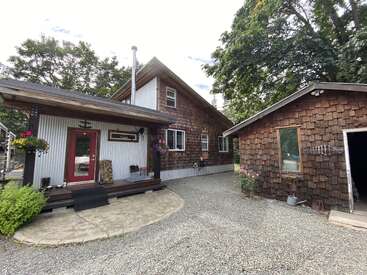 Ein charmantes, rustikales Haus mit Holzschindeln, einer einladenden roten Tür, Blumenkörben, einer Kiesauffahrt und einer separaten Garage, umgeben von Bäumen, viel Grün und einer friedlichen Atmosphäre.