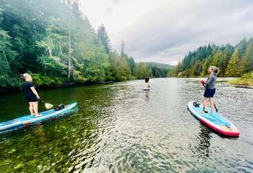 Drei Leute paddeln auf einem ruhigen Fluss, umgeben von dichtem, immergrünem Wald, über ihnen ein bewölkter Himmel, unter ihnen klares, seichtes Wasser – sie genießen gemeinsam ein friedliches, malerisches Abenteuer in der Natur.