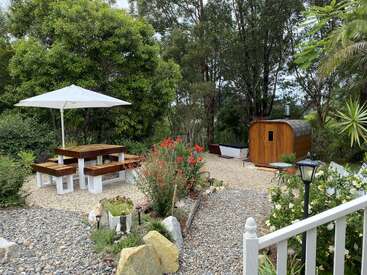 Uma cena tranquila de jardim com uma mesa de piquenique de madeira sob um guarda-sol branco, flores coloridas, vegetação exuberante, uma cabana de sauna de madeira e uma banheira ao ar livre.