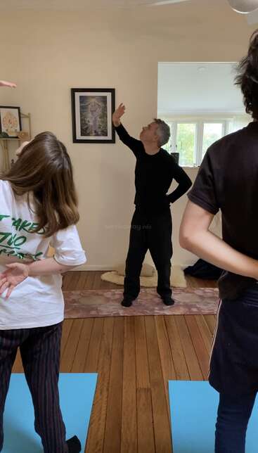 Três pessoas estão praticando ioga ou alongamento em um ambiente fechado. O instrutor, de preto, demonstra uma pose, enquanto dois participantes acompanham a atividade em tapetes azuis, no piso de madeira.