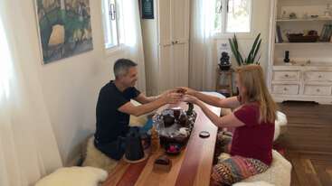 Um homem e uma mulher sentados em uma mesa de madeira, tomando chá em uma sala aconchegante e iluminada pelo sol, com cadeiras macias, plantas e decoração caseira. Atmosfera acolhedora.