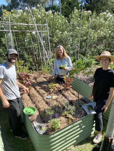 Three people are gardening together in a raised garden bed, surrounded by lush green plants and flowers, smiling and enjoying a sunny day outdoors.