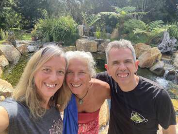 Three smiling adults pose together outdoors in front of a tranquil pond with rocks and lush greenery, enjoying a sunny day in a beautiful natural setting.