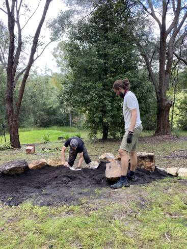 Deux personnes travaillent en plein air dans un jardin, s'affairant à disposer de gros rochers et de la terre. Elles sont entourées d'arbres et de verdure, ce qui crée un cadre paisible et naturel.