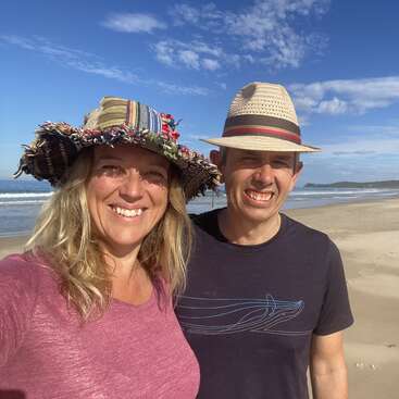 The image shows a man and woman standing on a beach, smiling at the camera, with the ocean and sky visible behind them.